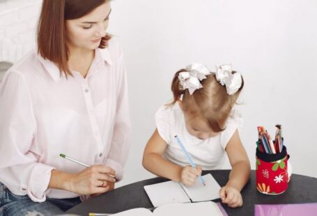 Childcare Schooling - From above small girl drawing with pencil in notepad while mother sitting near at round table