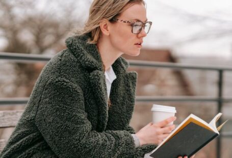 Winter Break - Side view of young female in warm clothes with takeaway coffee and open book sitting on bench in winter park